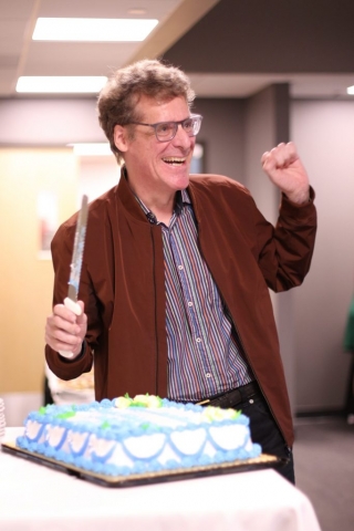 Jean Dufresne is standing by retirement cake in a celebratory pose. (Hands raised).