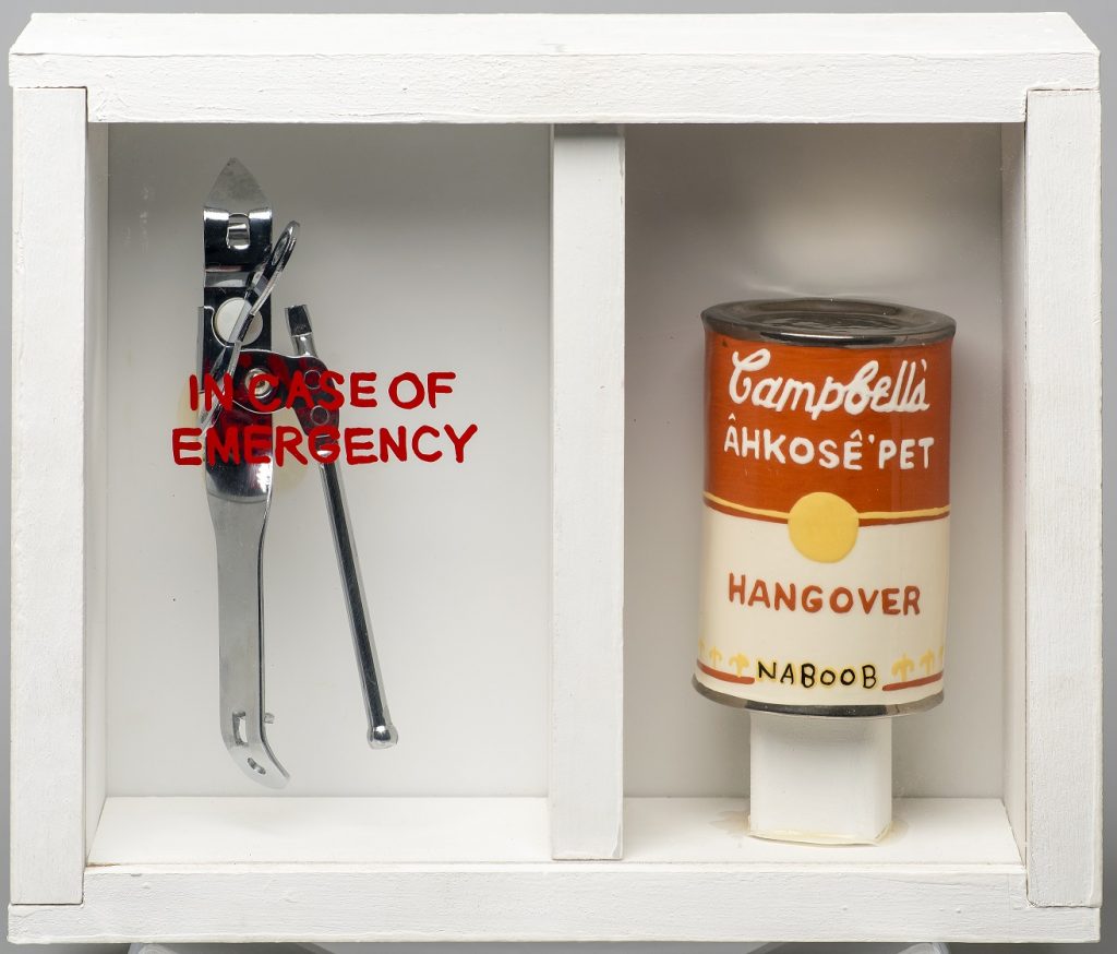 Image of a sculpture of a white, wooden box with a plexiglass front. On the left side is a silver can opener, underneath the words "In case of emergency" written on the glass. On the right side is a ceramic sculpture of a Campbell's soup can, with the words "CAMPBELL'S ÂHKOSÊ'PET / HANGOVER / NABOOB"