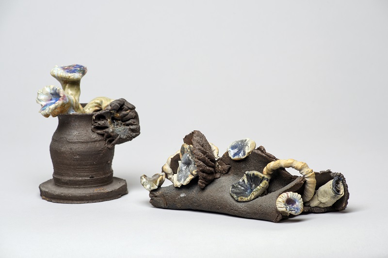 Image of two ceramic sculptures. On the left is an unglazed, brown vase with mushroom-like flowers emerging in light yellow and periwinkle. On the left, flowers grow like fungi from an unglazed, brown mass.