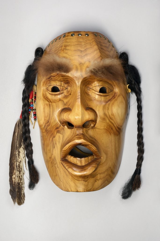 Image of a wooden mask with a surprised expression and an open mouth. The mask has furry eyebrows, black braids and a feather.