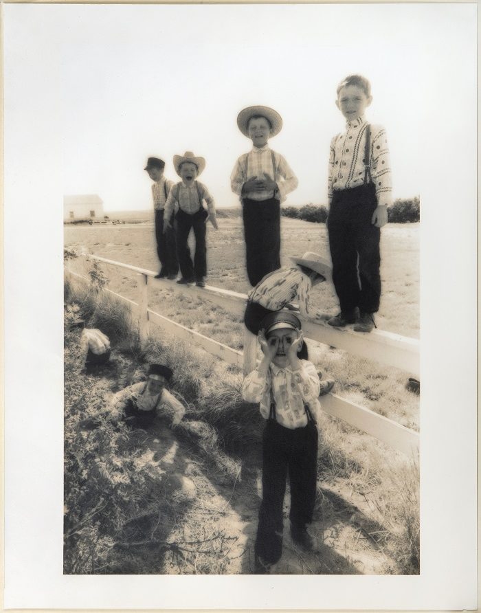 Soft-focus sepia toned photograph of a group of male children wearing traditional Hutterite clothing. They're standing in a field by a fence and making faces at the camera.